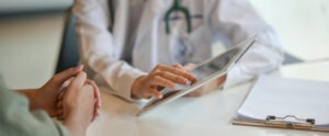 A doctor in a white coat with a stethoscope reviews information on a digital tablet with a patient seeking behavioral health in Nashville TN. A clipboard with paper is also on the desk.