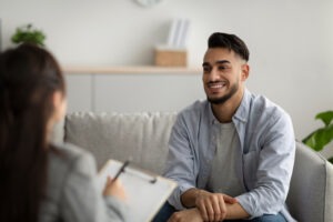 A man sits on a couch, smiling and talking to a woman with a clipboard, suggesting a counseling session at an addiction center in Nashville TN in a comfortable office setting.