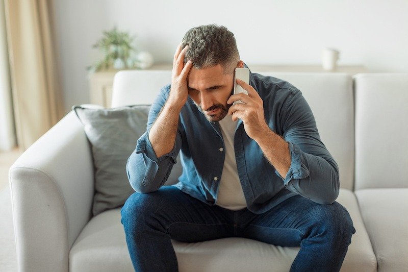 A worried man sits on a couch, holding a smartphone to his ear with one hand and resting his other hand on his head, appearing stressed or upset during a phone call about depression therapy in Nashville TN.