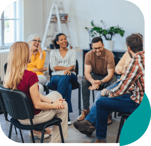 A diverse group of adults sits in a circle of chairs indoors, smiling and laughing during a casual group discussion at an addiction center in Nashville TN.