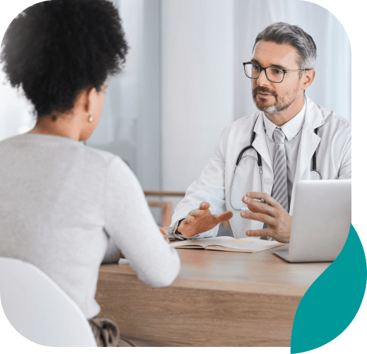 A doctor with glasses and a stethoscope talks to a patient across a desk in a medical office, discussing depression treatment in Nashville TN, with a laptop and an open notebook in front of them.