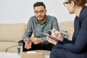 Two people sit on a beige couch having a serious conversation, possibly discussing behavioral health in Nashville TN. One gestures while talking, and the other takes notes with a clipboard. A glass of water and a box are on the table between them.