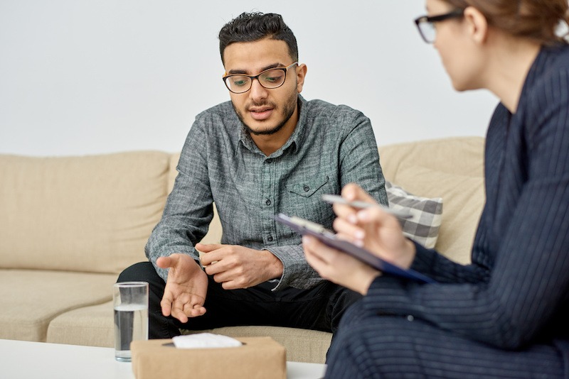 Two people sit on a beige couch having a serious conversation, possibly discussing behavioral health in Nashville TN. One gestures while talking, and the other takes notes with a clipboard. A glass of water and a box are on the table between them.