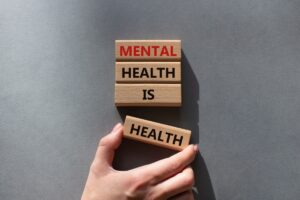 A hand places a wooden block labeled "HEALTH" next to three blocks stacked to read "MENTAL HEALTH IS HEALTH" on a gray background, highlighting the importance of mental well-being and depression treatment in Nashville TN.