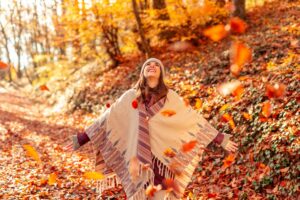 A woman wearing a poncho and beanie stands in a forest covered with autumn leaves, smiling joyfully as she tosses colorful leaves into the air—her happiness reflecting the hope found at a leading addiction center in Nashville TN.