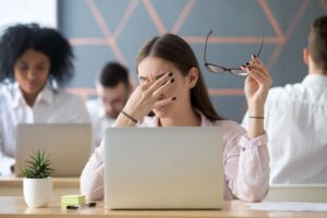 A woman sits at a desk holding her glasses and rubbing her eyes in front of a laptop, appearing tired or stressed—perhaps considering seeking behavioral health in Nashville TN. Other people are working in the background.