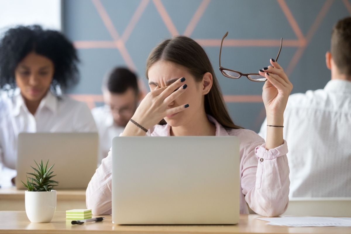 A woman sits at a desk holding her glasses and rubbing her eyes in front of a laptop, appearing tired or stressed—perhaps considering seeking behavioral health in Nashville TN. Other people are working in the background.
