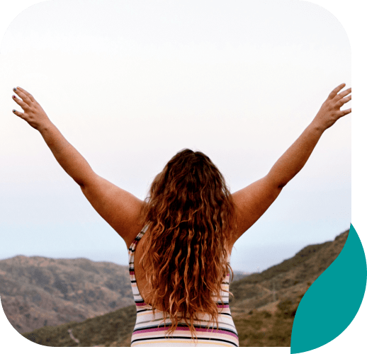 A person with long wavy hair raises their arms in the air while standing outdoors, facing distant hills under a pale sky. The person is wearing a sleeveless striped top.