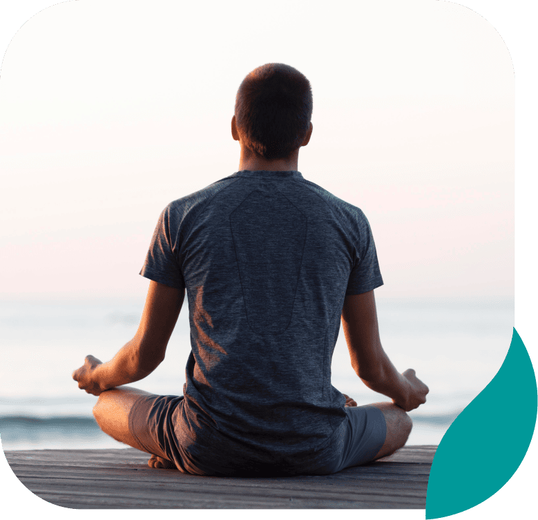 A person in a gray shirt sits cross-legged on a wooden surface facing the ocean, meditating at sunrise or sunset, with a calm and peaceful atmosphere.