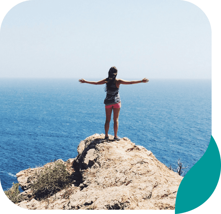 A person stands on a rocky cliff with arms outstretched, overlooking a vast, calm blue ocean under a clear sky. Some greenery is visible on the rocks in the foreground.