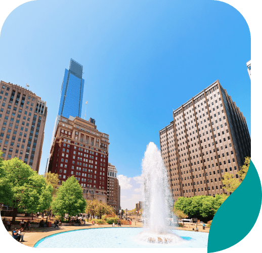 A city park with a large water fountain in the center, surrounded by green trees and tall buildings under a clear blue sky.