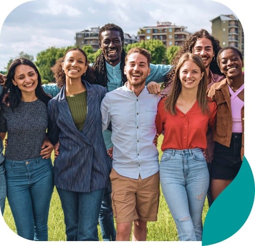 A diverse group of young adults stands close together outdoors, smiling and posing for the camera on a sunny day, with green grass and apartment buildings in the background.