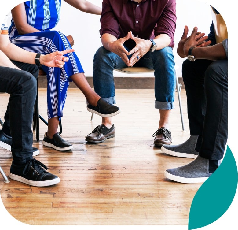 Four people sit in a circle on chairs, engaged in conversation. Only their lower bodies are visible. The floor is wooden, and the atmosphere appears casual and informal.