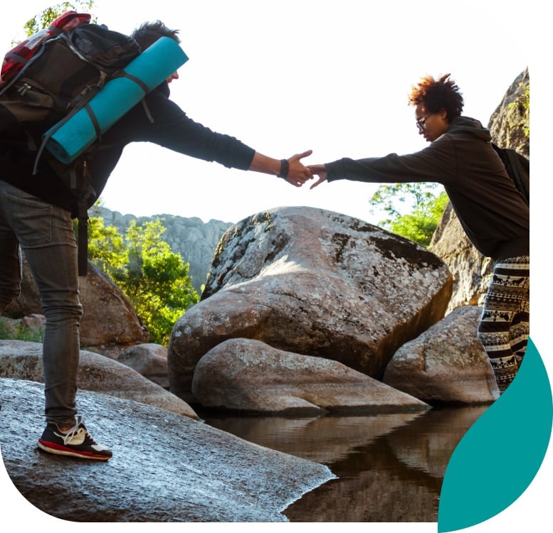 Two hikers reach out and hold hands while crossing rocks over a stream in a mountainous, scenic outdoor setting, surrounded by large boulders and greenery under a clear sky.