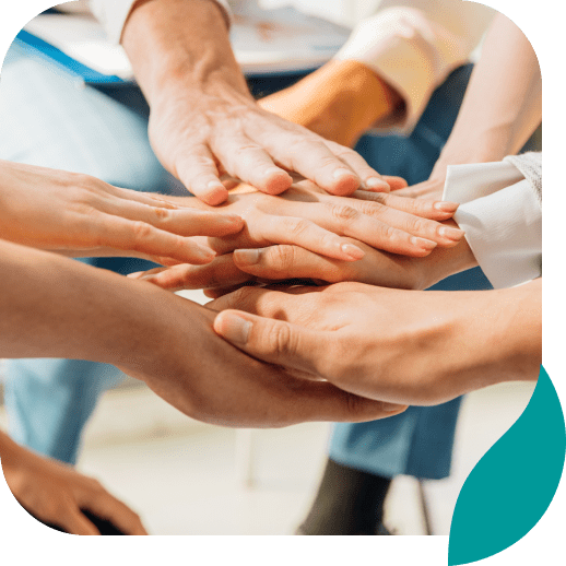 Several people place their hands on top of each other in a teamwork gesture, symbolizing unity and collaboration. The background is blurred, focusing on the hands.
