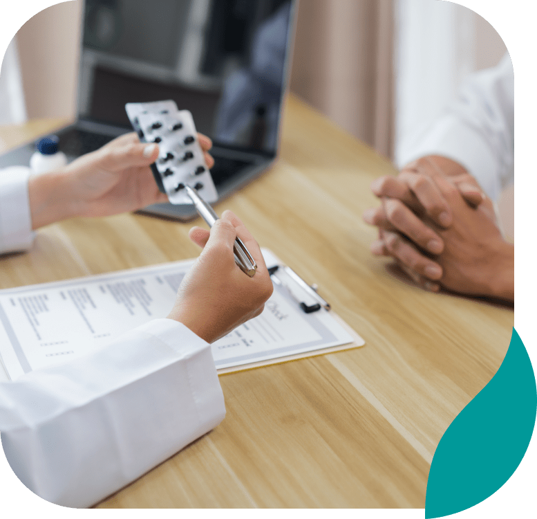 A doctor holding a blister pack of pills and a pen discusses medication with a patient at a desk with a clipboard and laptop.