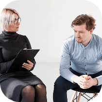 A woman with glasses and a clipboard sits facing a man who appears pensive and is looking down, with his hands clasped. They are seated in a bright room, suggesting a counseling or therapy session.