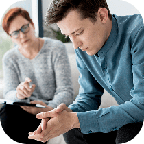 A man sits with his head down, looking troubled, while a woman with glasses and a clipboard listens attentively, suggesting a therapy or counseling session.