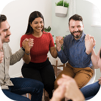 A group of people sitting in a circle, smiling and holding hands, appearing to participate in a supportive group activity or discussion in a bright, indoor setting.