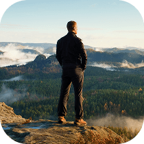 A person stands on a rocky cliff edge, looking out over a vast forested landscape with rolling hills and mist rising in the distance under a clear sky.