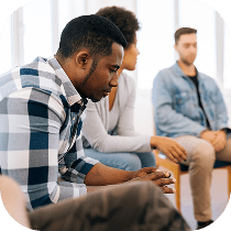 A man in a plaid shirt sits pensively in a support group setting, with two other people sitting nearby, all appearing thoughtful and engaged in conversation.