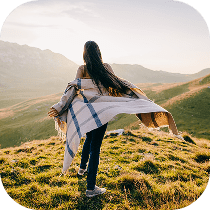 A woman with long hair stands on a grassy hilltop, facing away, holding a light-colored blanket. The sun is setting over distant mountains, casting a warm glow over the landscape.