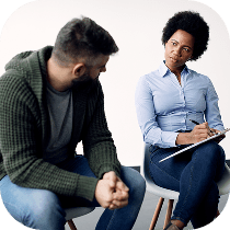 A man sits with his hands clasped, looking at a woman who is writing on a clipboard. The woman appears to be listening attentively, suggesting a counseling or therapy session. Both are seated on chairs against a plain background.