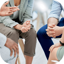 Four people sit in a close circle, engaged in conversation. Only their hands and arms are visible, with gestures indicating discussion or support, suggesting a group meeting or therapy session.