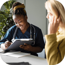 A healthcare professional in navy scrubs, wearing a stethoscope, smiles while writing on a clipboard and talking to a patient seated across the desk.