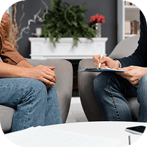 Two people sit across from each other in chairs; one is holding a clipboard and writing, the other has hands clasped. A white table and potted plants are in the background, suggesting a therapy or counseling session.