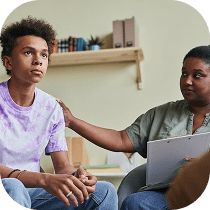 A concerned young person sits with hands clasped while an adult with a clipboard gently rests a hand on their shoulder, offering support during a conversation in a comfortable, home-like setting.