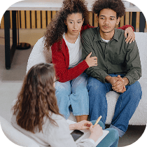 A couple sits closely together on a couch, looking concerned, while talking to a therapist who is taking notes during a counseling session.