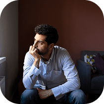 A man with dark curly hair and a beard sits indoors, resting his chin on his hand, gazing thoughtfully out of a window. He wears a light blue shirt and appears deep in thought.