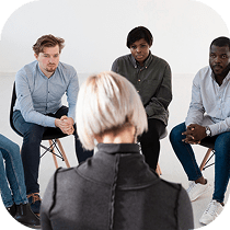 A group of four people sit in chairs in a circle, attentively listening to a person with short blond hair who faces them, suggesting a support or discussion group setting.