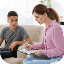 A teenage boy sits on a couch while a woman in a pink blouse writes on a clipboard, possibly during a counseling or therapy session in a bright, comfortable room.