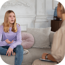 A teenage girl with long blonde hair, wearing a lavender sweater and jeans, sits on a sofa talking to an adult who is taking notes, suggesting a therapy or counseling session.