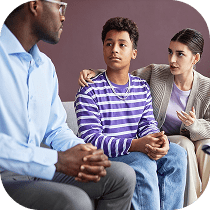 A teenage boy in a purple striped shirt sits between a woman with her arm around him and a man in glasses, all engaged in a serious conversation.