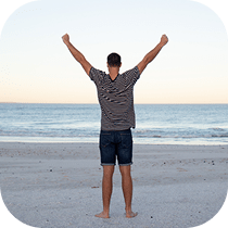 A person stands on a sandy beach facing the ocean with arms raised triumphantly in the air. The sky is clear and calm, creating a peaceful, uplifting scene.