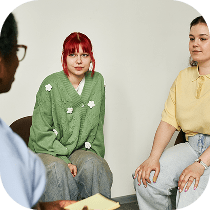 Three people sit in a circle having a discussion; one person with red hair and a green sweater faces forward, another in a yellow top sits nearby, and a third person with a notepad is partially visible in the foreground.