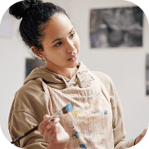 A woman wearing a paint-stained apron and hoodie holds a paintbrush, looking thoughtfully to the side in an art studio with artwork on the walls behind her.