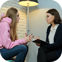 A young woman in a pink hoodie speaks seriously with a female therapist, who is holding a clipboard and pen, in a well-lit room with a lamp and light-colored walls.