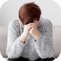 A person with brown hair sits on a couch, leaning forward with their head down and hands clasped in front of their face, appearing stressed or upset. They are wearing a gray sweater.
