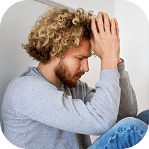 A man with curly blond hair and a beard sits with his head bowed and hands pressed together on his forehead, appearing stressed or deep in thought. He wears a light gray sweater and blue jeans.