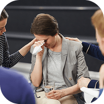 A woman sitting and wiping her eyes with a tissue appears upset while others around her offer comfort by gently touching her shoulders and arms.