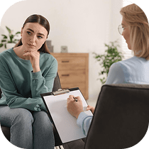 A woman sits thoughtfully with her hand on her chin while talking to a therapist, who is taking notes on a clipboard during a counseling session in a comfortable, well-lit room.
