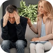 A concerned woman gently touches the shoulder of a distressed man sitting next to her on a sofa, as he holds his head in his hands. Both appear to be indoors, with a leafy plant in the background.