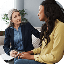 A therapist with gray hair listens attentively and comforts a young woman in a yellow shirt during a counseling session on a couch.