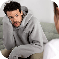 A man in a gray hoodie sits on a sofa, leaning his head on his hand and looking concerned, while facing another person whose back is visible in the foreground.
