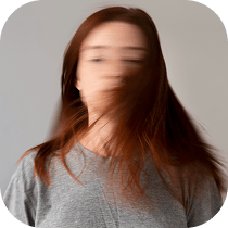 A person with long brown hair wearing a gray shirt, facing the camera with their head moving quickly, causing their face and hair to appear blurred. The background is plain light gray.