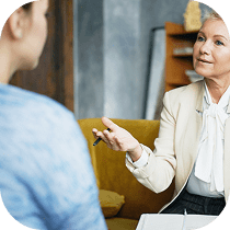 An older woman in a light blazer speaks attentively to another person, who is facing away from the camera, in an indoor setting, possibly during a counseling or consultation session.